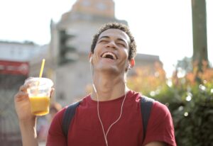 A smiling young man in a red shirt listens to music and holds a beverage outdoors, expressing joy.